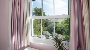 A room with a large window, white curtains, and a view of a white fence and greenery outside.