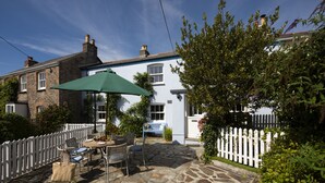 A stone patio with outdoor furniture and a green umbrella, surrounded by a white picket fence and greenery.