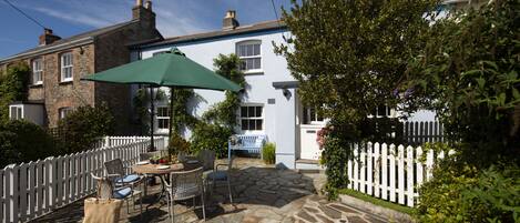 A stone patio with outdoor furniture and a green umbrella, surrounded by a white picket fence and greenery.