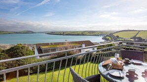 Un balcon avec une table dressée pour le dîner, offrant une vue sur un paysage côtier avec une étendue d’eau et des collines lointaines.