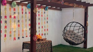 Un espace patio agrémenté d’un fauteuil suspendu, d’une petite table recouverte d’une nappe à motifs floraux et de décorations en papier colorées accrochées au mur.