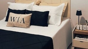 Un lit bien fait, recouvert d’une couvre-lit noire et blanche, avec un oreiller beige orné des lettres « WIA » et un oreiller noir. On aperçoit une table de chevet avec une lampe et une petite boîte en bois portant des chiffres.