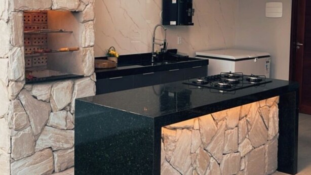 Une cuisine avec un mur d’accent en pierre, des plans de travail noirs, une cuisinière intégrée et un meuble noir au-dessus de l’évier.