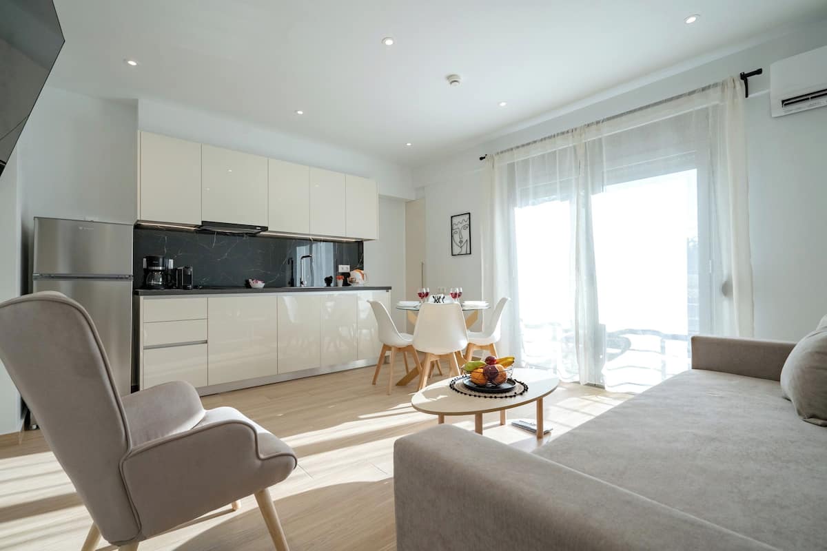 Uma sala de estar moderna com sofá e poltrona, uma área de jantar com mesa e cadeiras, uma cozinha com armários brancos e bancada preta, e janelas amplas com cortinas leves.