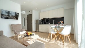 A modern living room with a sofa, a dining table with chairs, and a kitchen area with cabinets and appliances. - Arte 80 (Spata-Artemida)