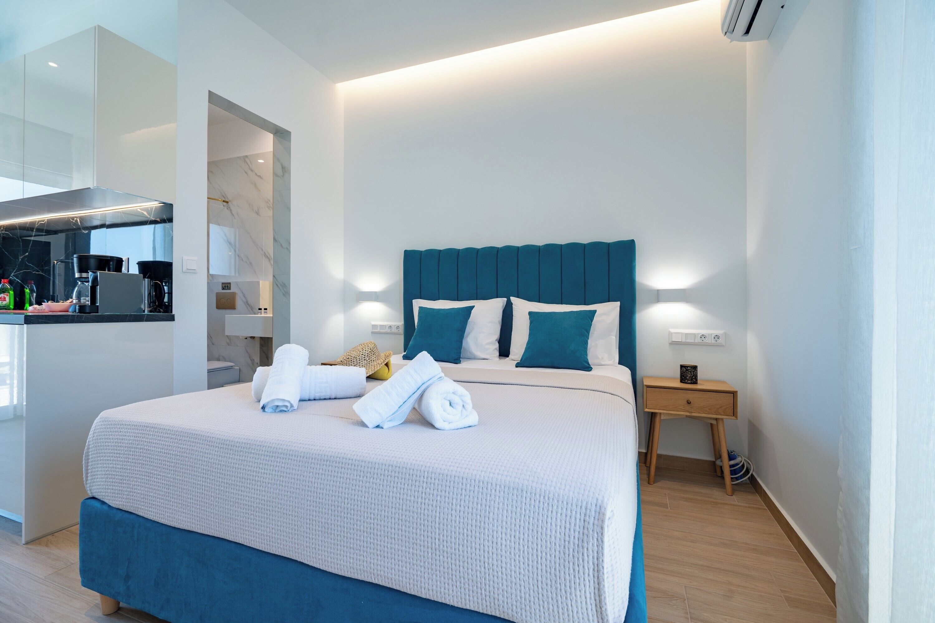 A modern hotel room with a large bed, a bedside table, a small desk, and a built-in kitchen area.