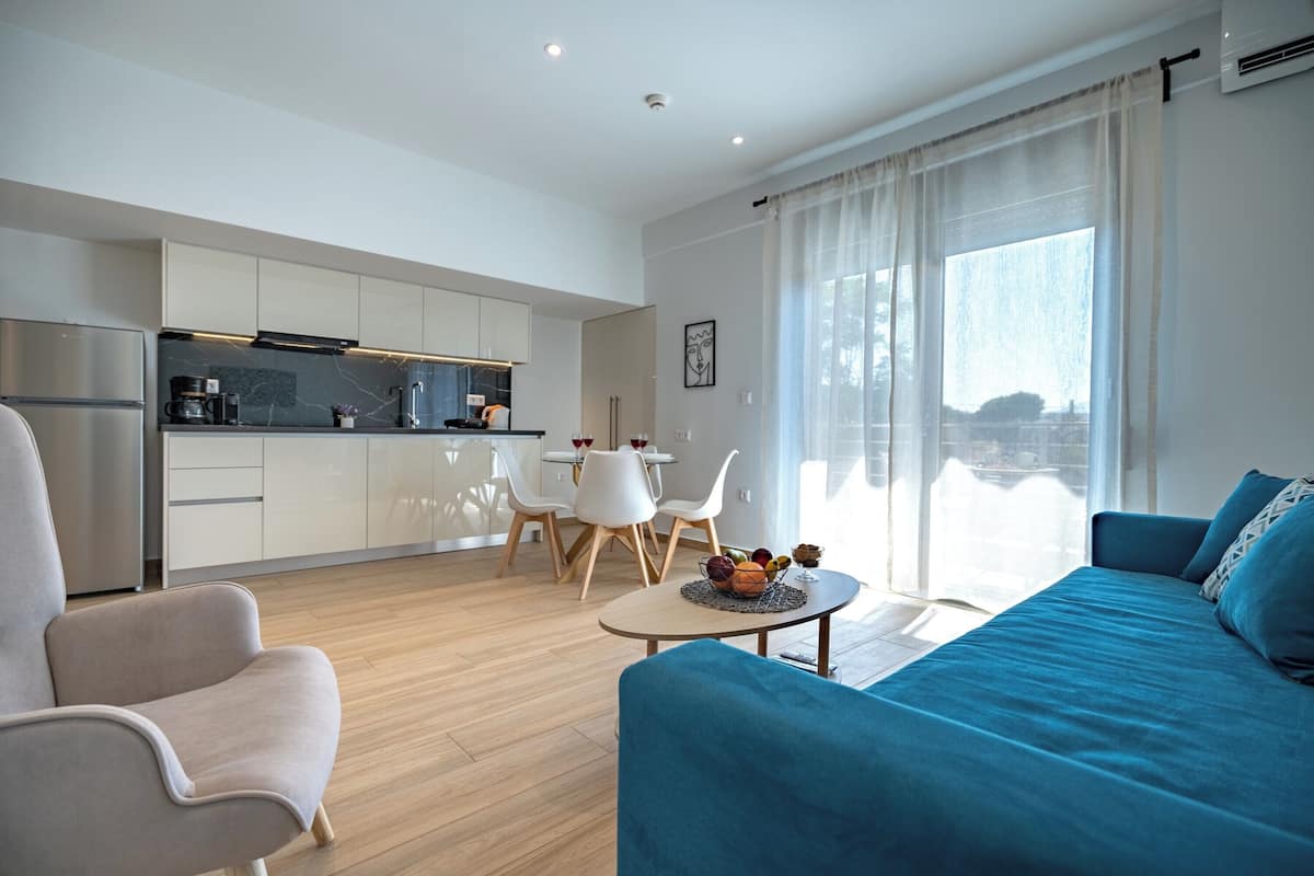 Uma sala de estar moderna com um sofá azul, uma poltrona clara e um espaço de jantar com uma mesa e cadeiras brancas.