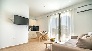 A modern living room with a sofa, a dining area with a table and chairs, a flat-screen TV mounted on the wall, and a kitchen in the background. - Arte 80 (Spata-Artemida)