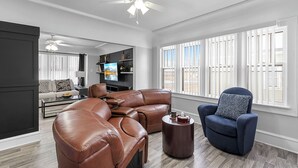 A spacious living room with a large brown leather sectional sofa, a round coffee table, and a blue armchair. There is a ceiling fan, a flat-screen TV, and a dining area visible in the background.