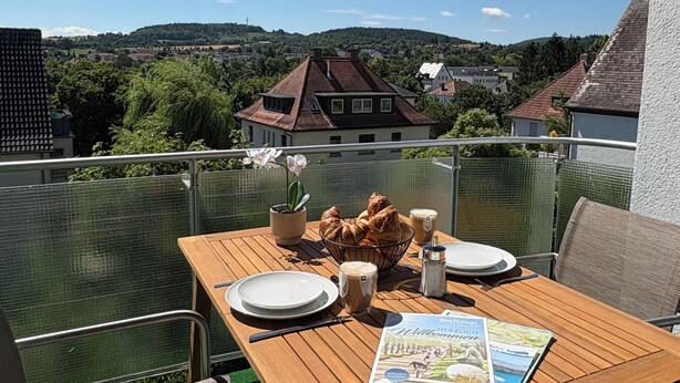 Mesa de madera dispuesta para dos personas, con platos, una taza de café y un menú en un balcón con vistas a una zona residencial con casas y vegetación.
