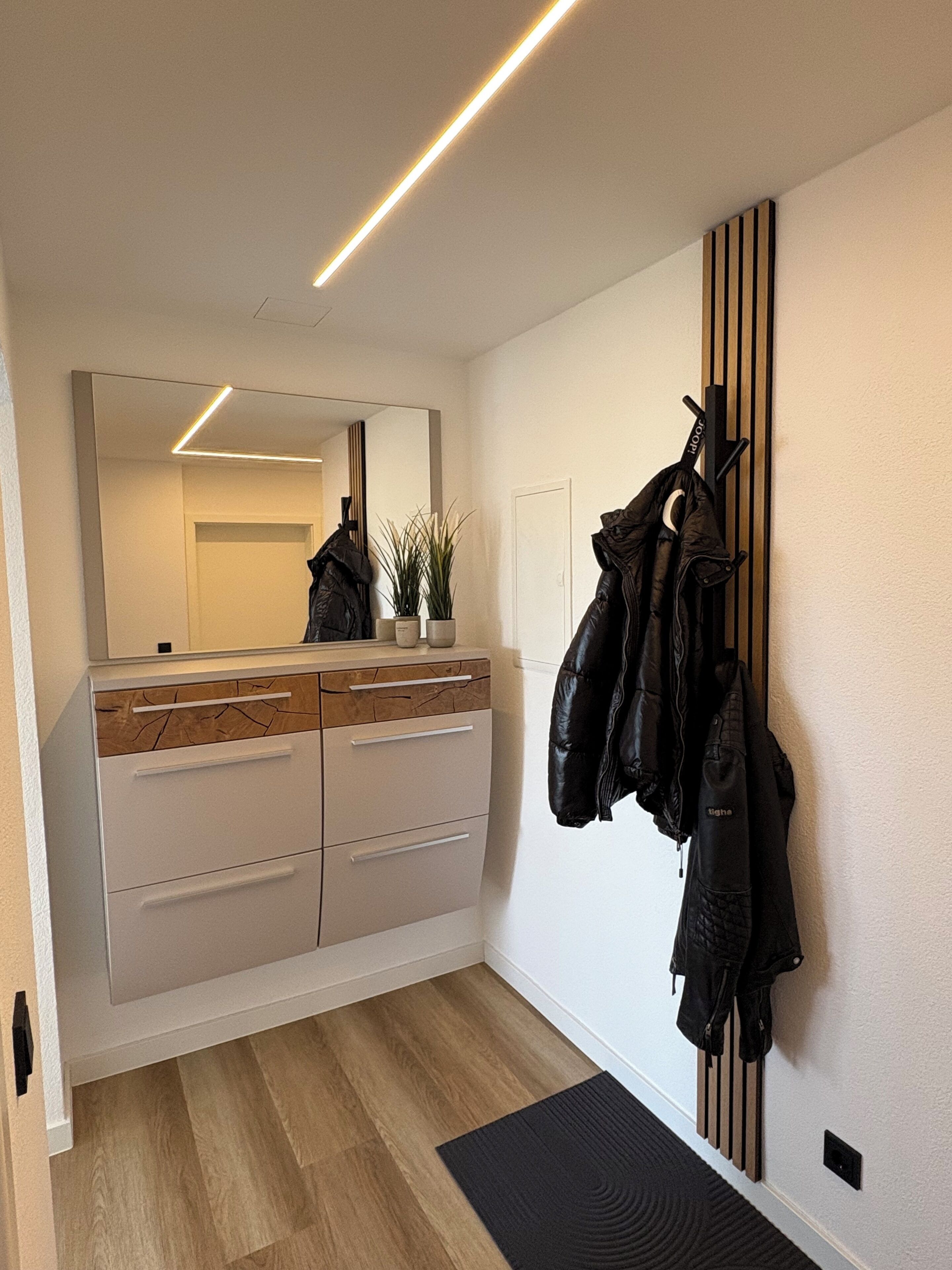 A modern hallway with a wooden floor, a white cabinet with drawers, a mirror, and a coat rack with jackets.