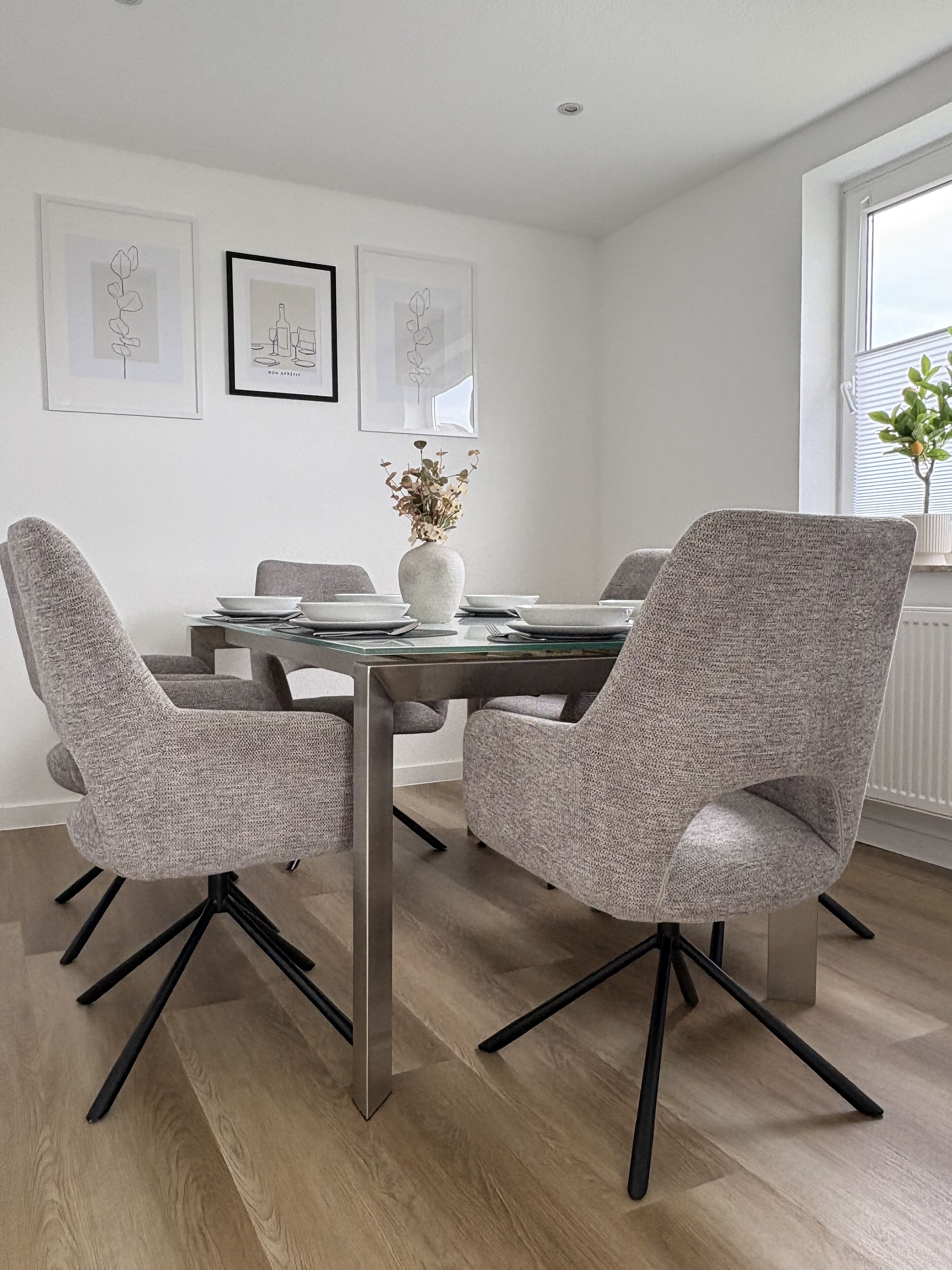 Ein moderner Essbereich mit einem Tisch aus Glasplatte, grau gepolsterten Stühlen und einer Vase mit getrockneten Blumen.