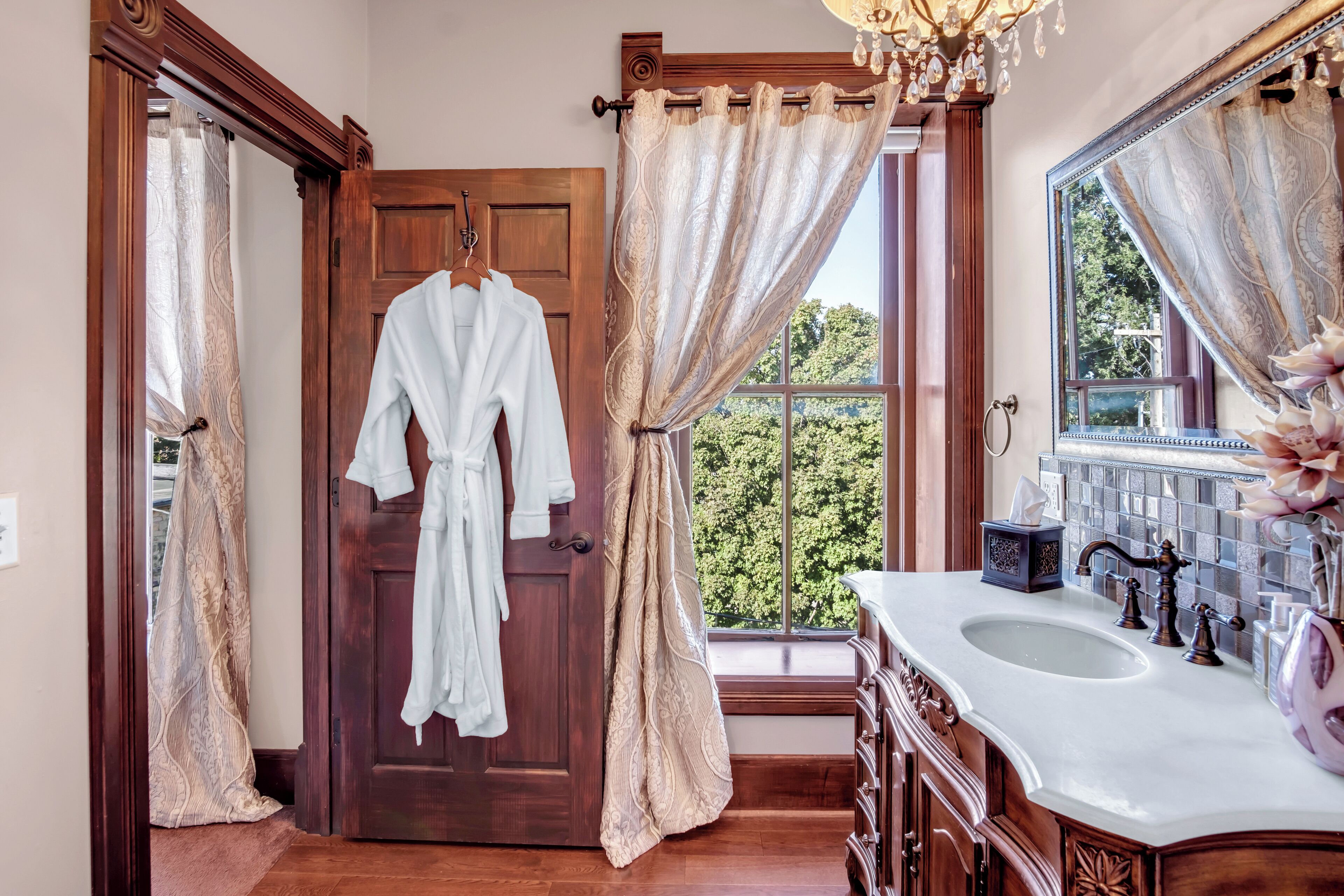 Une salle de bain avec une porte en bois, un peignoir blanc accroché à un porte-manteau, un grand miroir et un lavabo agrémenté d’un vase décoratif.