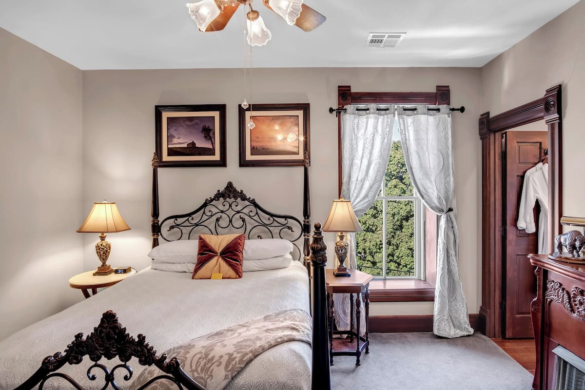 A bedroom with a bed, two bedside tables with lamps, a ceiling fan, a window with curtains, and a door with a coat rack.