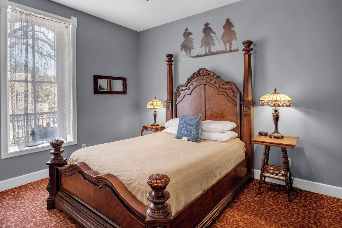 A bedroom with a large bed, two bedside tables with lamps, a window with sheer curtains, and a wall mural.