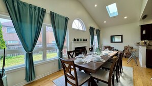 Uma sala de jantar com uma mesa comprida posta para seis pessoas, grandes janelas com cortinas, uma lareira e uma área de cozinha.