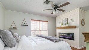 A bedroom with a bed, a ceiling fan, a fireplace, and a window with blinds.
