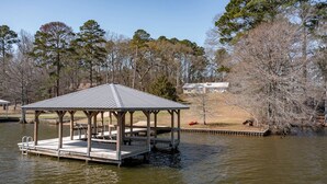 A wooden gazebo on a lake with a dock, surrounded by trees and a distant house.