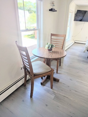 Uma área de refeições com mesa redonda e duas cadeiras de madeira, um pequeno vaso com flores e vista para o verde lá fora.