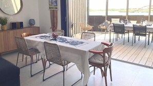 A dining area with a table set for four, surrounded by wicker chairs. A large mirror hangs on the wall, and a poster is visible. The room has a view of the outdoors through large windows.