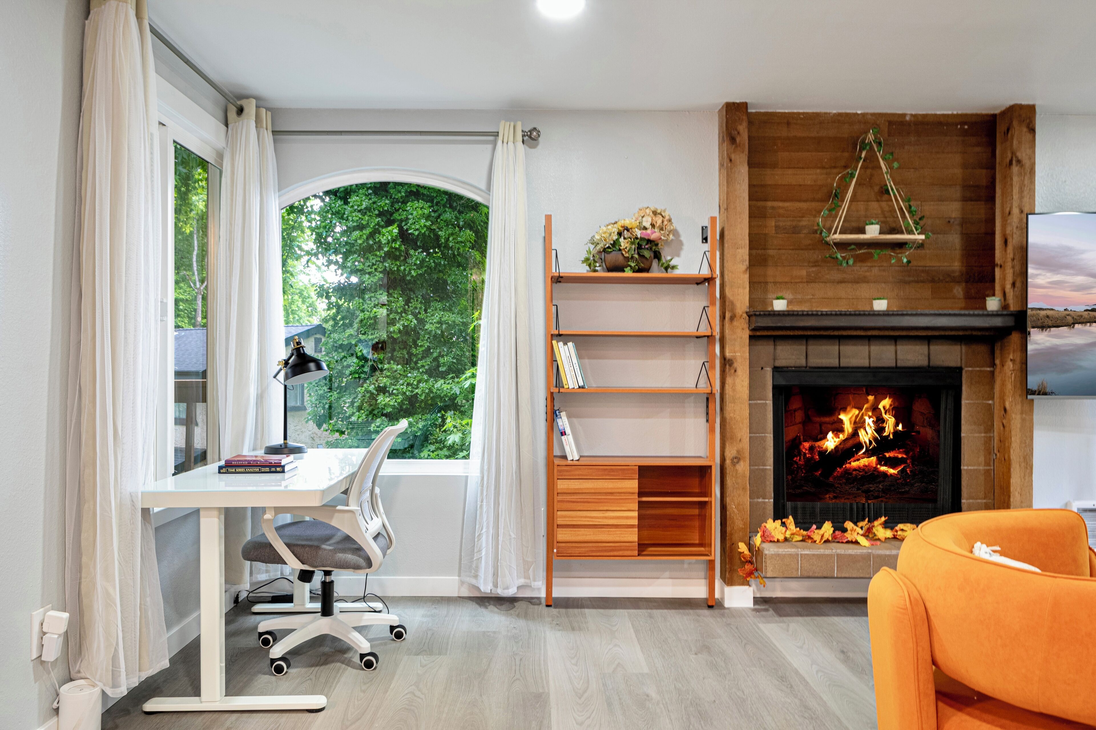 A room with a desk, chair, bookshelf, fireplace, and a large window with curtains.