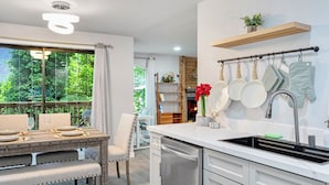 Une cuisine moderne dotée d’une grande fenêtre, d’un coin repas avec une table dressée pour deux personnes et d’un porte-ustensiles suspendu.