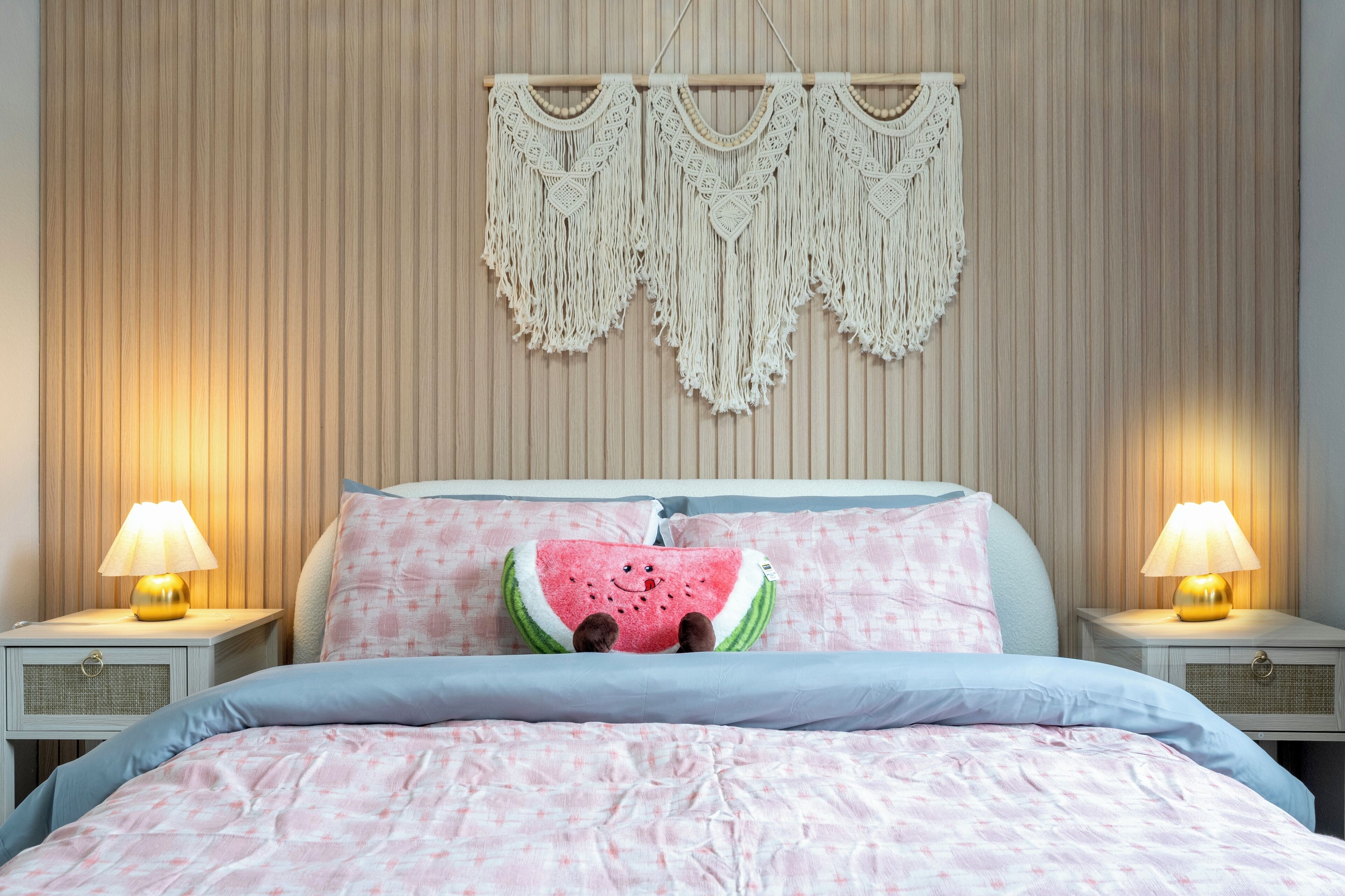 Una cama con una colcha estampada en rosa y blanco, un cojín con forma de sandía y un tapiz de macramé colgado en la pared.