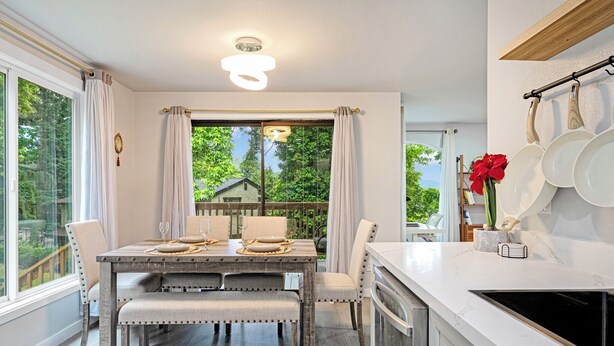 Un coin repas avec une table en bois dressée pour quatre personnes, une cuisine avec des plans de travail blancs et une vue sur la verdure à l’extérieur.