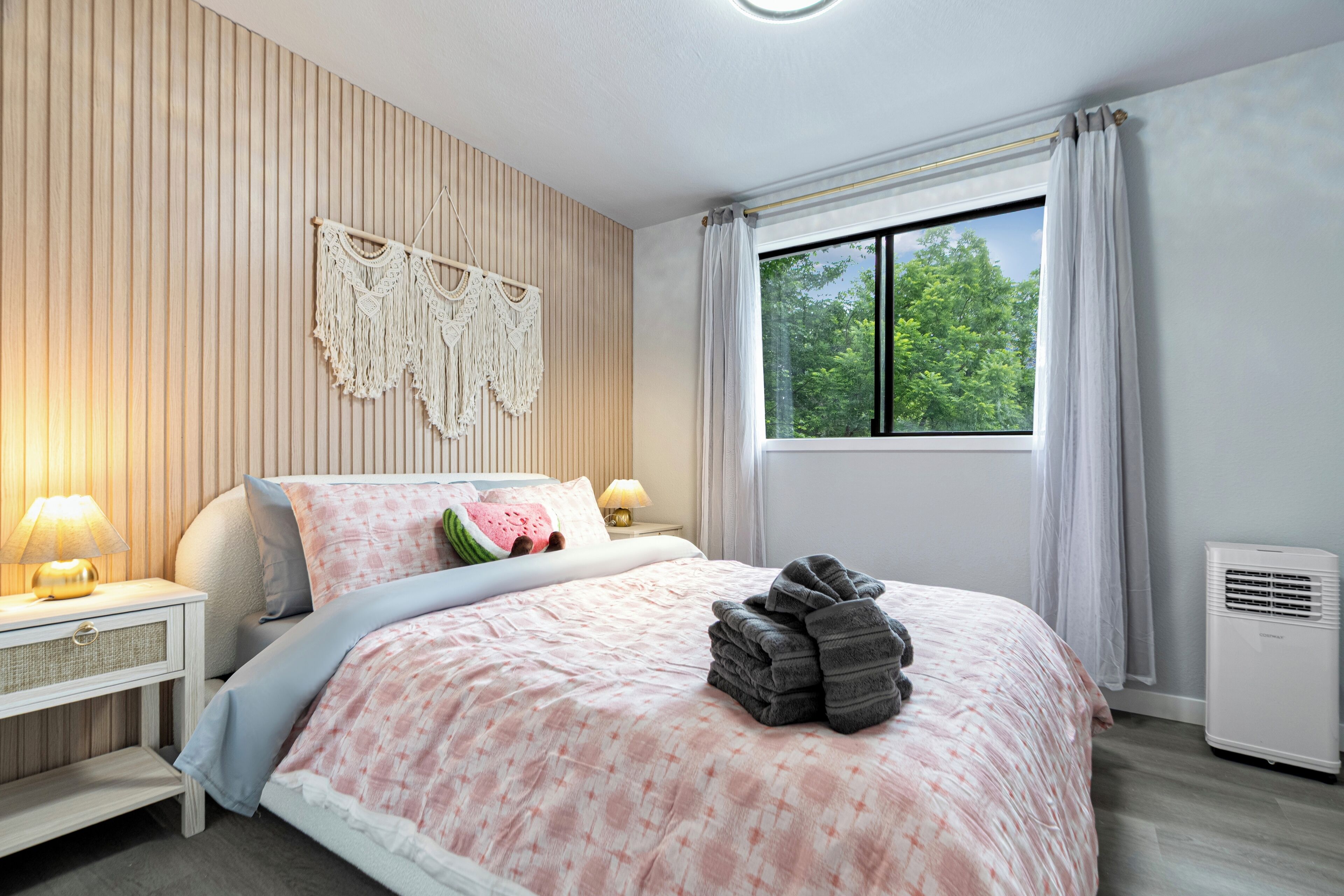 Ein Schlafzimmer mit Bett, Nachttischlampen, einer Wanddeko aus Makramee, einem Fenster mit Vorhängen und einer Heizung.