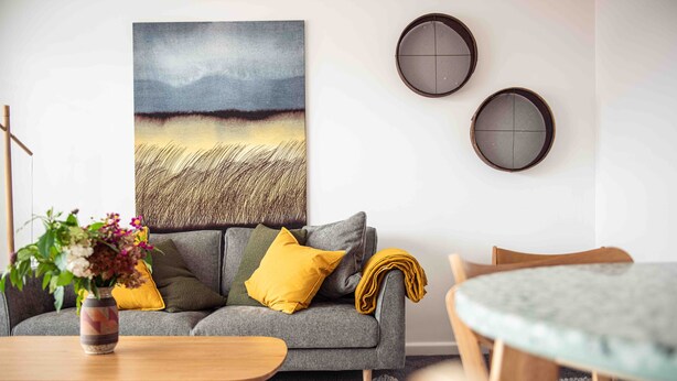 A living room with a grey sofa, a wooden coffee table, and a painting of a field. There are two round wall decorations and a vase with flowers on the coffee table.