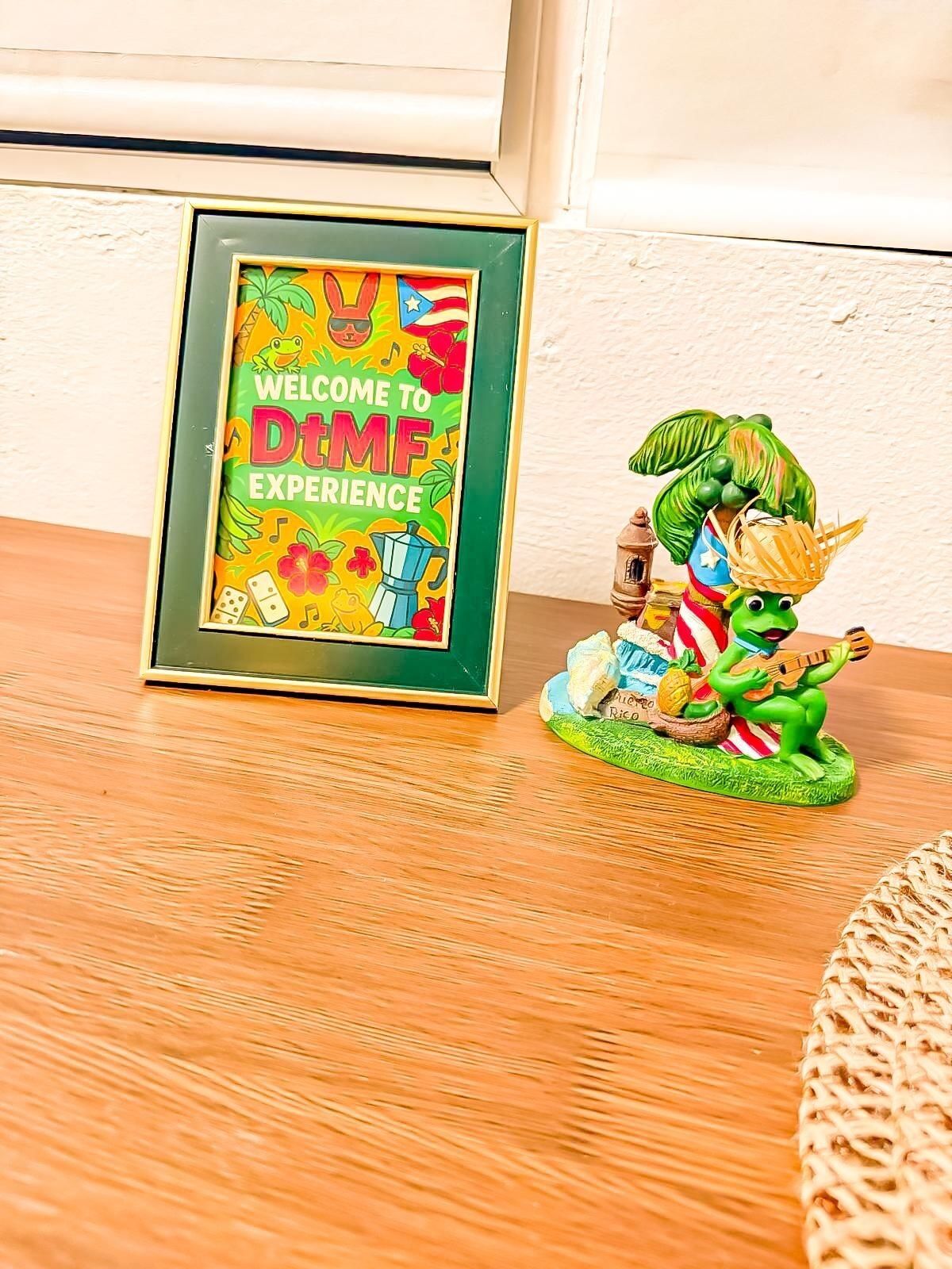 Un letrero de bienvenida enmarco con motivos tropicales y una pequeña figurita de un sapo verde tocando la guitarra sobre una superficie de madera.