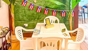 A dining area with a green backdrop, white plastic chairs, and a table with a menu and a small plant. - DtMF Experience at his Hometown in Vega Baja - Near Beach and Natural Springs! (Vega Baja)