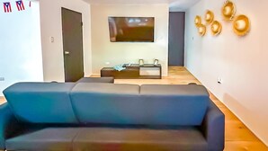 A modern living room with a large black sofa, a flat-screen TV mounted on the wall, and a wooden floor. - DtMF Experience at his Hometown in Vega Baja - Near Beach and Natural Springs! (Vega Baja)
