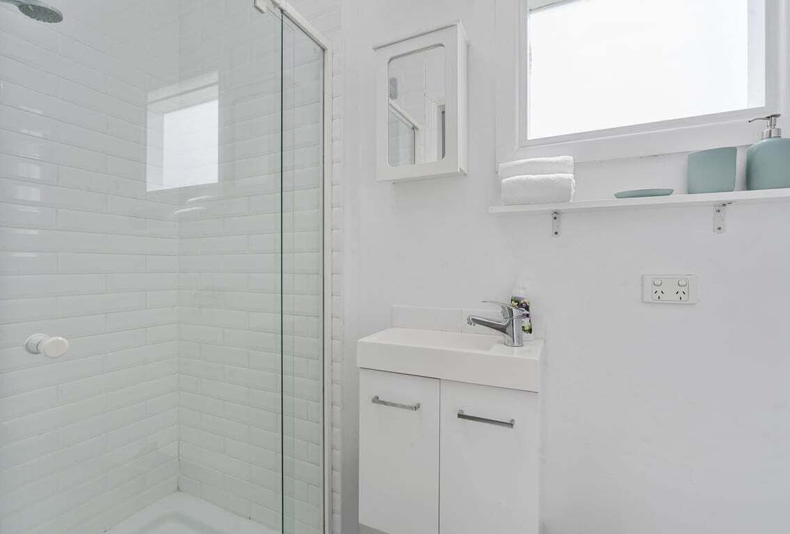 A bathroom with white tiled walls, a glass shower enclosure, a white sink with a mirror above it, and a small window.