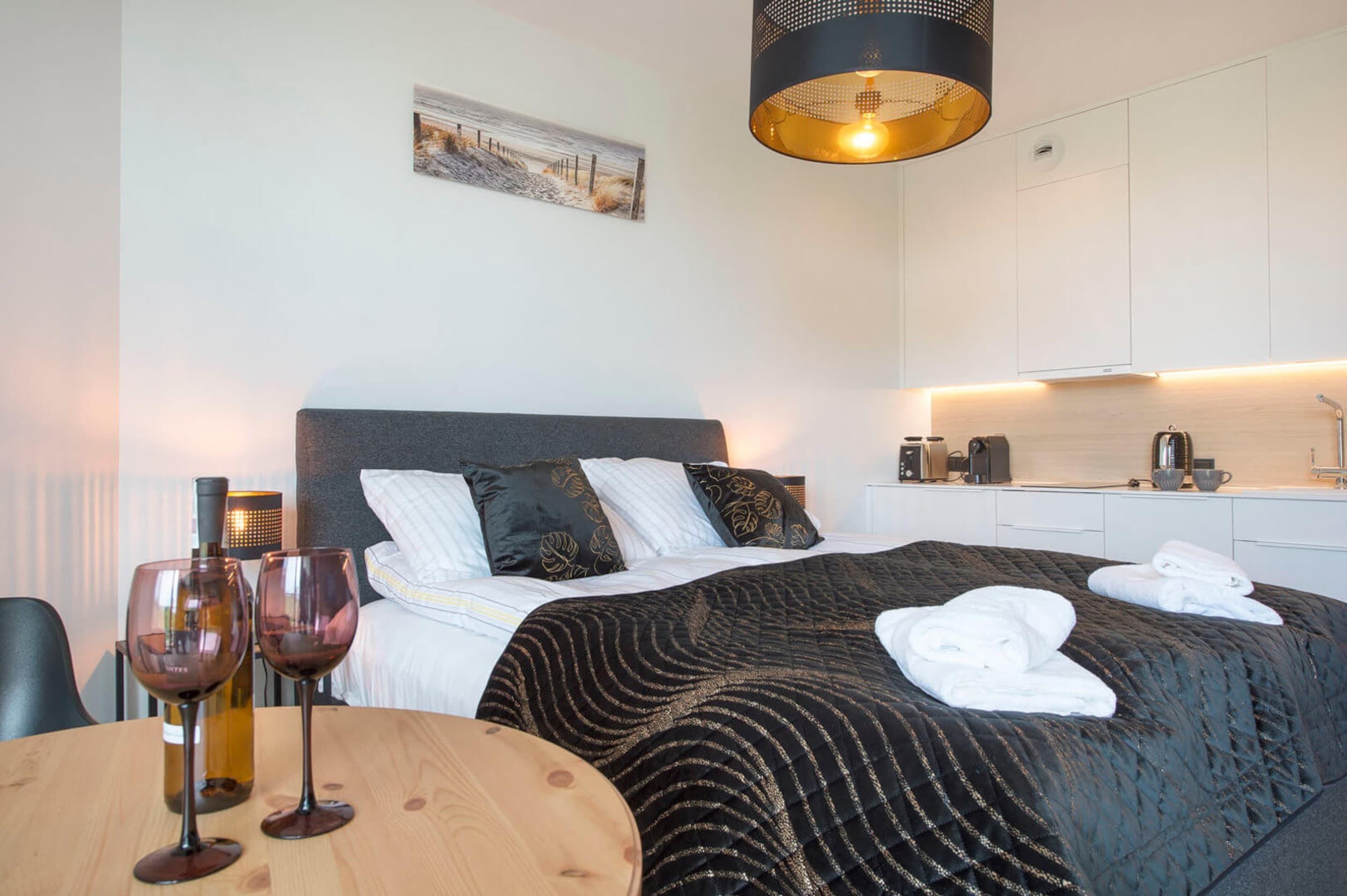 Una habitación de hotel con una cama, una mesita de noche con copas de vino, una mesa redonda de madera, un cuadro en la pared y una cocina moderna.