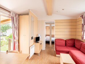 A compact room with a wooden floor, a red sofa, a small table, a TV, and a window with curtains. - Mobile Home in Loire With Pool (Veuzain-sur-Loire)