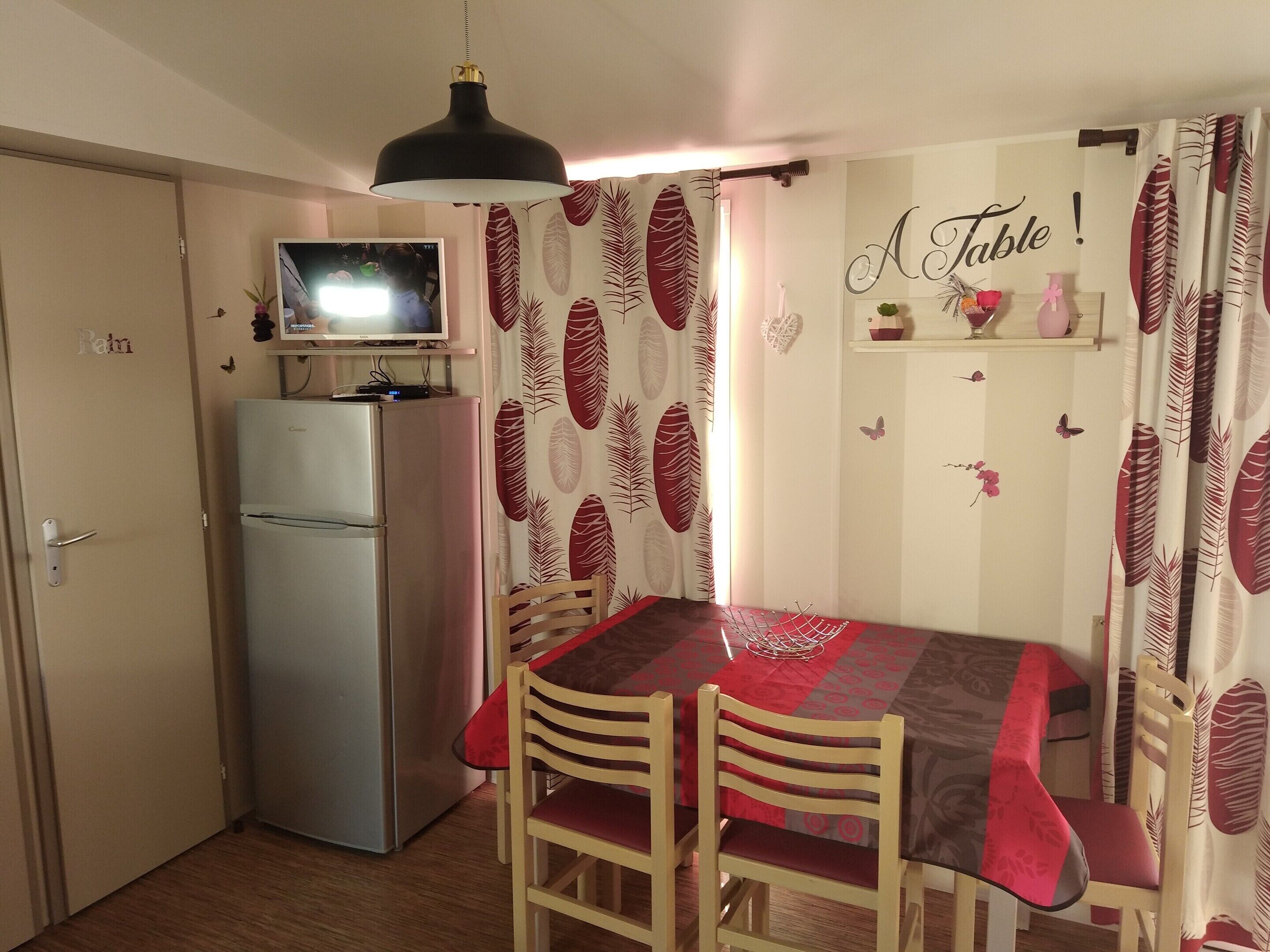 A compact kitchen with a refrigerator, a dining table with chairs, a television above the fridge, and a wall with a 'A Table!' sign.