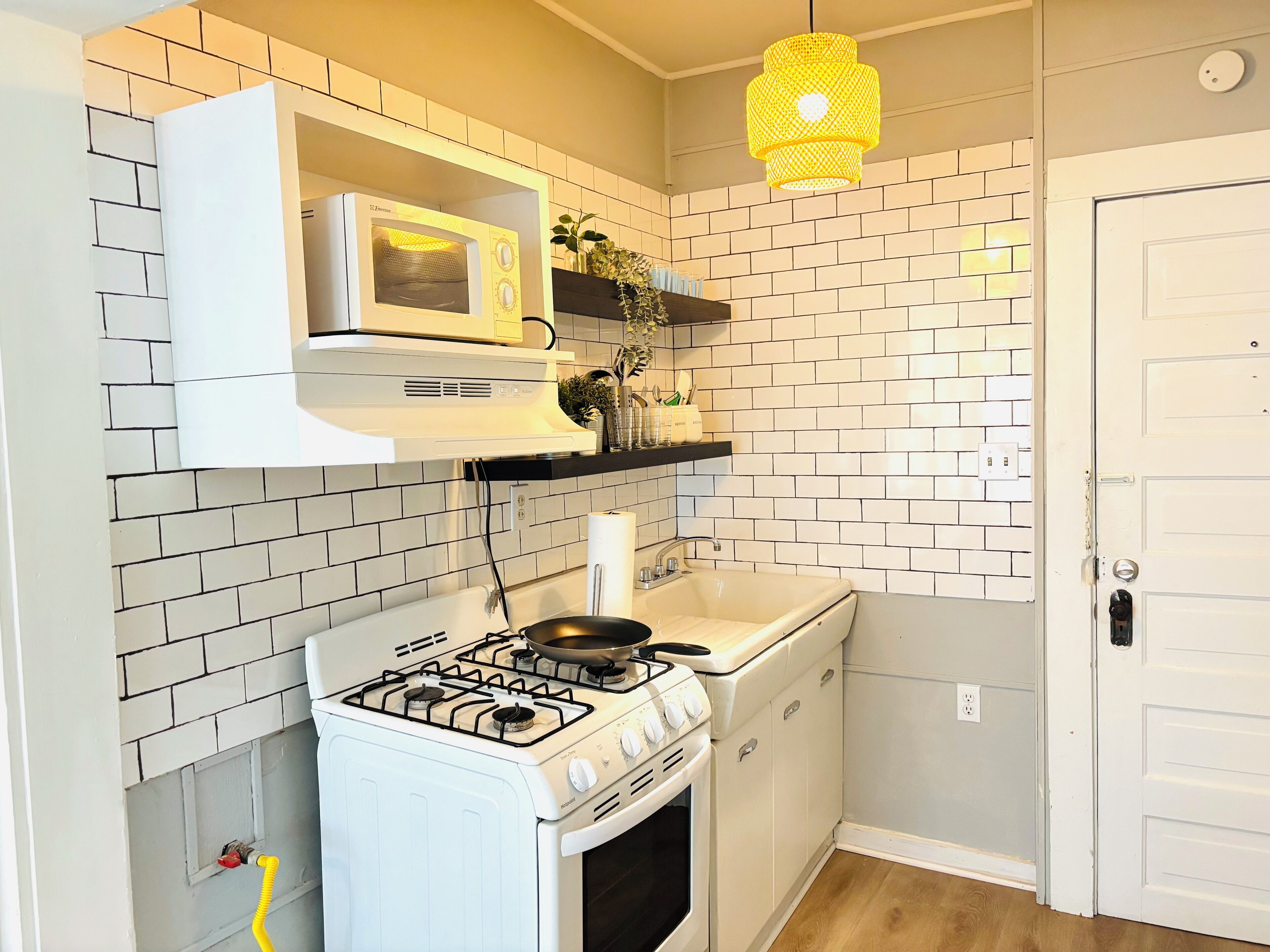 Et lille køkken med gasblus, mikroovn og vask. Der er hvide skabe og en gul hængende lampe.