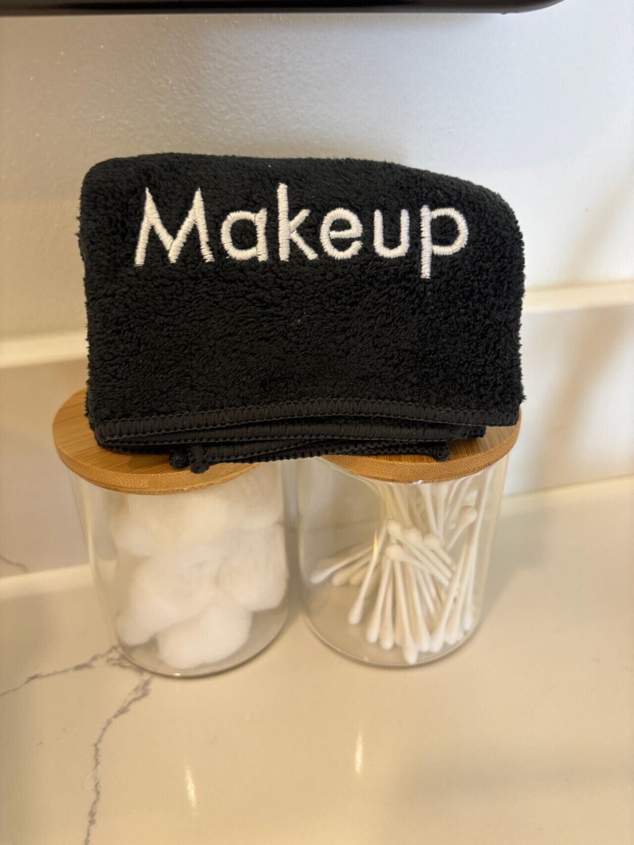 Una toalla negra con el texto 'Makeup' bordado, sobre un soporte de madera encima de un frasco de vidrio con bastoncillos de algodón.