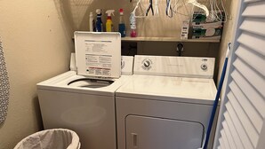 Un espace buanderie comprenant un lave-linge et un sèche-linge à chargement frontal, un panier à linge en osier et des produits de nettoyage rangés sur une étagère.