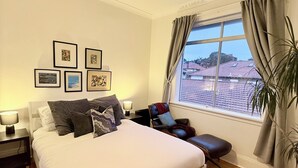 Quarto com cama, ventilador de teto, janela com cortinas, mesa de cabeceira e quadros na parede.