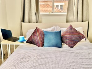 A bed with a laptop on a small table beside it, a cup, and a window with curtains.