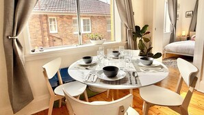 Uma sala de jantar com mesa redonda posta para quatro pessoas, cadeiras brancas e vista para um edifício de tijolos através de uma janela.