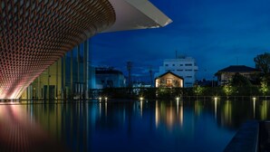 A modern building with a unique roof structure, a reflective pool, and a small house in the background.