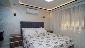 A modern bedroom with a large bed, a nightstand, a wall-mounted air conditioner, and a window with sheer curtains.