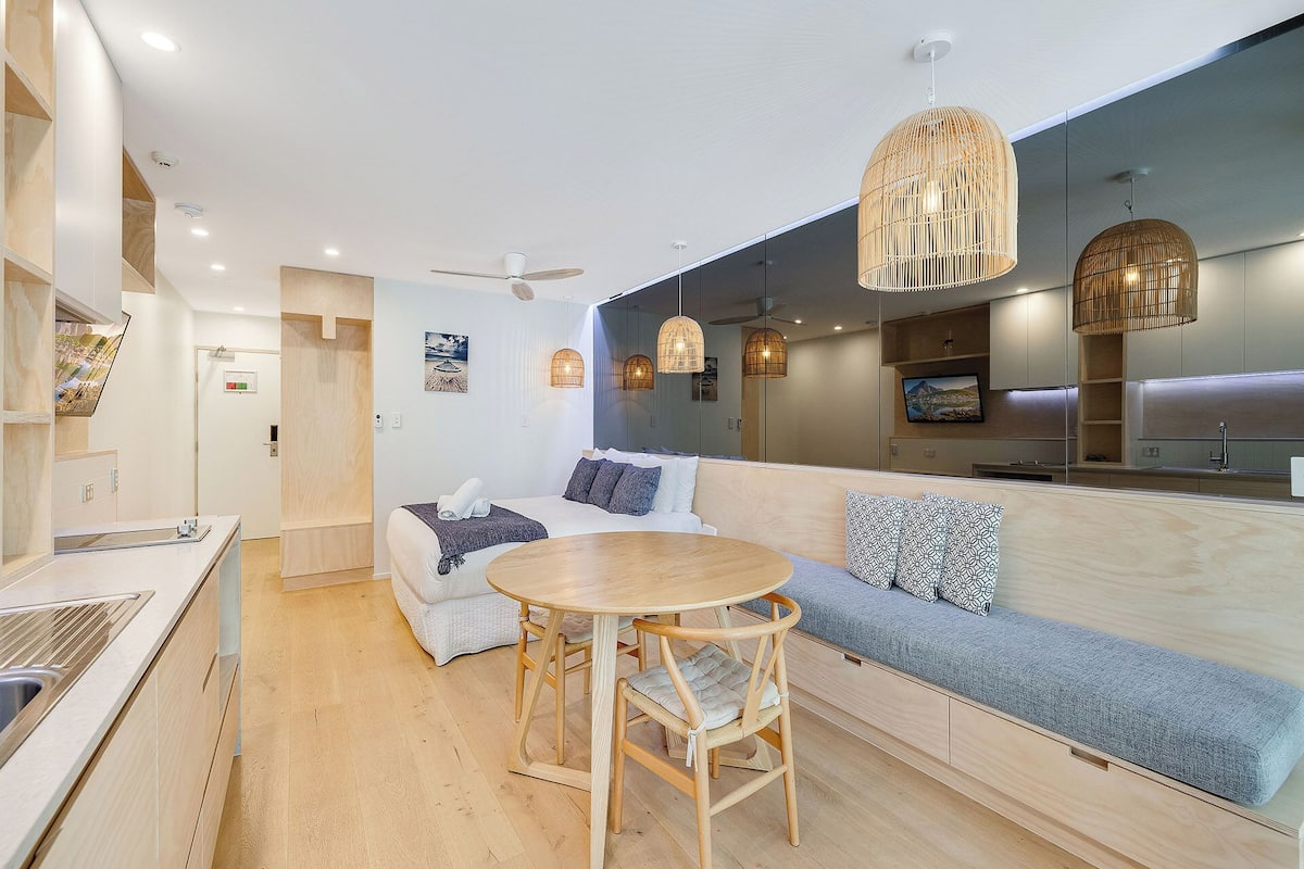 A modern hotel room with a kitchenette, a round dining table with chairs, a sofa with cushions, and a large mirror.