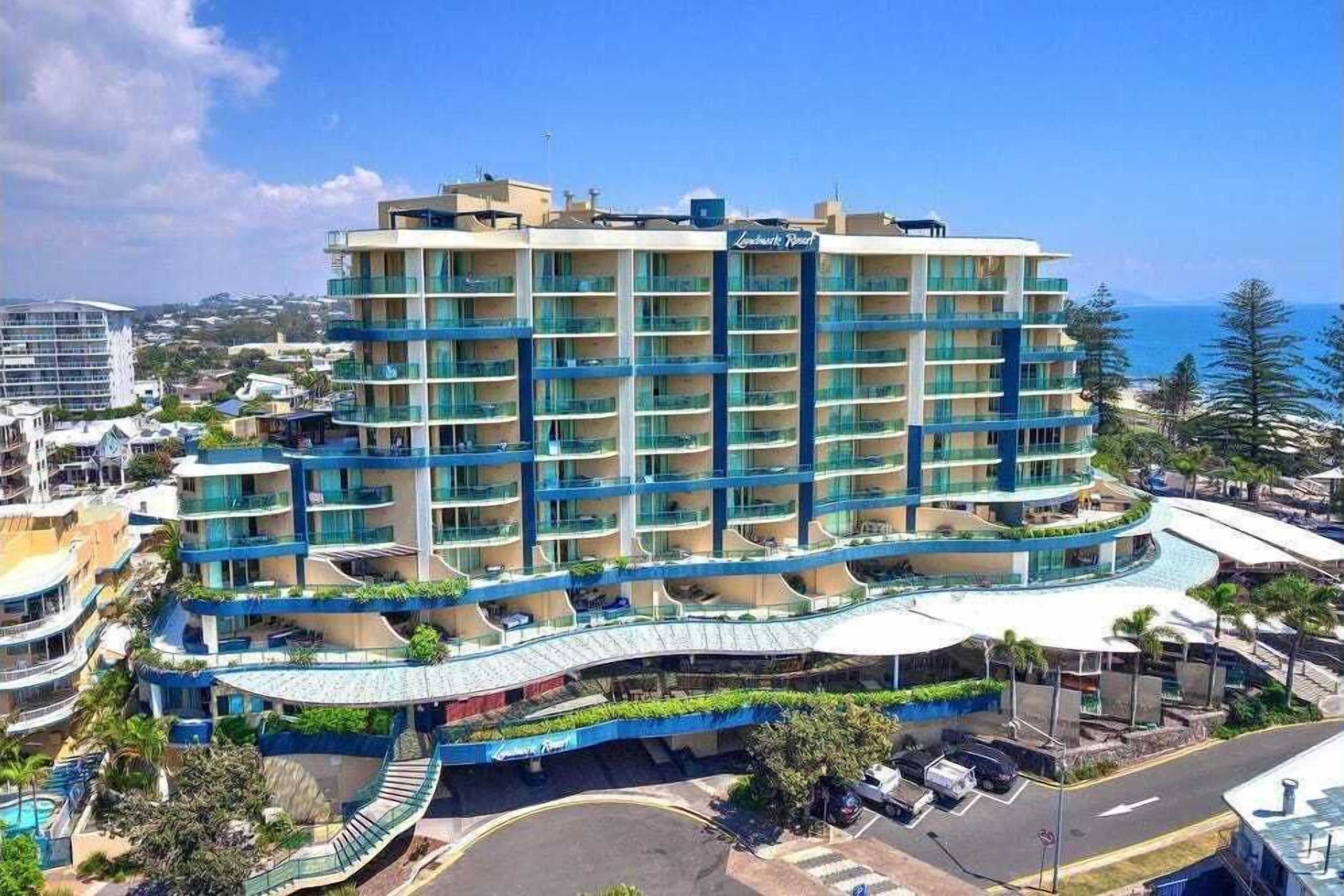 A multi-story hotel with a curved balcony, located near a beach with clear skies.