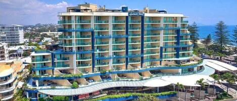 A multi-story hotel with a curved balcony, located near a beach with clear skies.