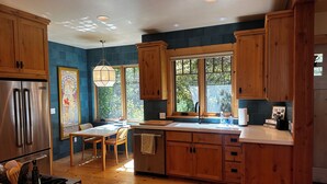 A modern kitchen with wooden cabinets, a stainless steel refrigerator, a dining area with a table and chairs, and a large window providing natural light. - Walk to Downtown Graeagle, Mill Pond & Pickleball (Graeagle)