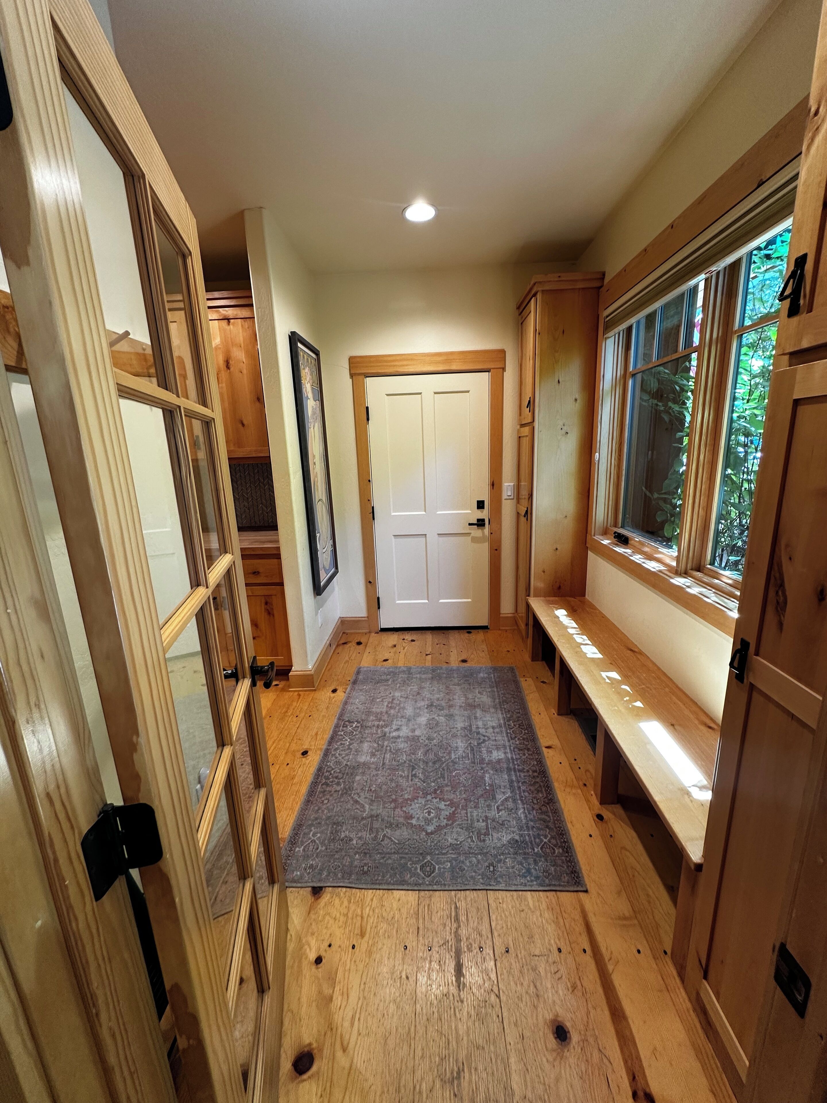 Un couloir doté d’un plancher en bois, d’un tapis gris, d’un banc, d’une porte et d’une fenêtre donnant sur un paysage verdoyant.